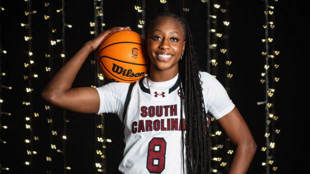 Joyce Edwards in white uniform with basketball on her shoulder in front of white background with small white W shaped lights behind her.